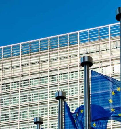 Drei Flaggen der Europäischen Union wehen vor einem modernen Glasgebäude mit einer gitterartigen Fassade unter einem klaren blauen Himmel.