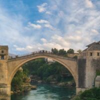 Eine historische Steinbogenbrücke überspannt einen Fluss und verbindet mittelalterliche Gebäude mit Steinmauern und Ziegeldächern, umgeben von üppigem Grün und Bergen unter einem teilweise bewölkten Himmel.