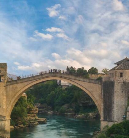 Eine historische Steinbogenbrücke überspannt einen Fluss und verbindet mittelalterliche Gebäude mit Steinmauern und Ziegeldächern, umgeben von üppigem Grün und Bergen unter einem teilweise bewölkten Himmel.