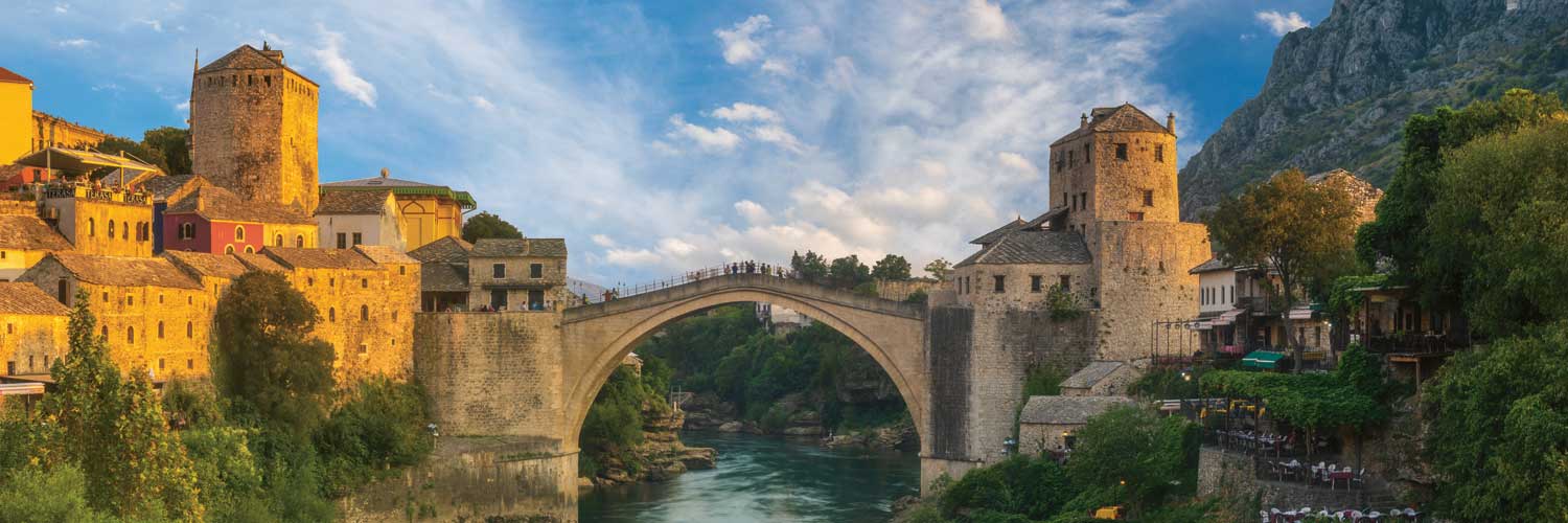 Eine historische Steinbogenbrücke überspannt einen Fluss und verbindet mittelalterliche Gebäude mit Steinmauern und Ziegeldächern, umgeben von üppigem Grün und Bergen unter einem teilweise bewölkten Himmel.