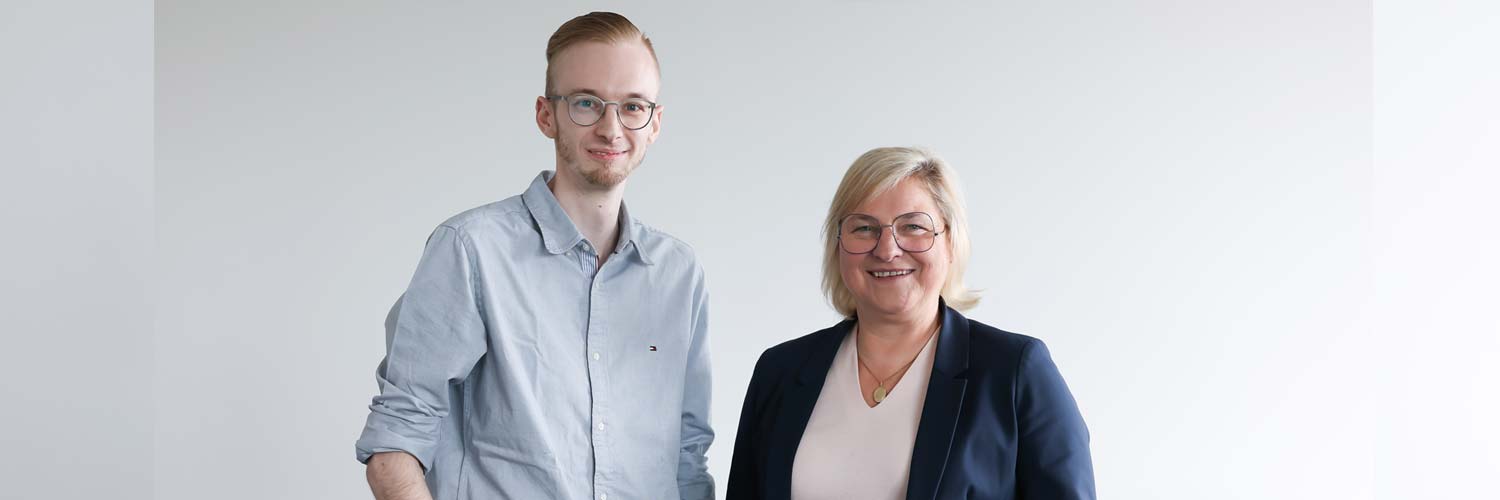 Ein junger Mann in einem hellblauen Hemd und einer Brille steht neben einer älteren Frau in einem marineblauen Blazer und einer Brille. Beide lächeln und schauen in die Kamera vor einem schlichten hellen Hintergrund.