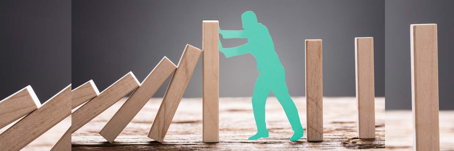 Die blaugrüne Silhouette einer Person stützt einen Holzklotz und verhindert, dass eine Reihe fallender Dominosteine die verbleibenden aufrechten Klötze auf einer Holzfläche umstürzt.