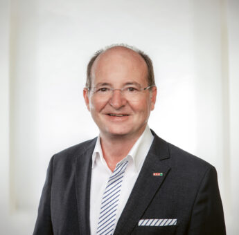 Ein Mann mittleren Alters im Anzug und mit gestreifter Krawatte steht lächelnd vor einem hellen, verschwommenen Hintergrund. Er trägt eine Brille und hat eine kleine Anstecknadel mit roten und grünen Farben an seinem Revers.