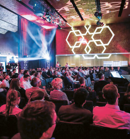 Ein großes Publikum sitzt in einem modernen, schwach beleuchteten Auditorium und verfolgt eine Präsentation auf der Bühne. Helle bunte Lichter und geometrische Muster schmücken die Decke und die Wände und schaffen eine lebendige Atmosphäre.