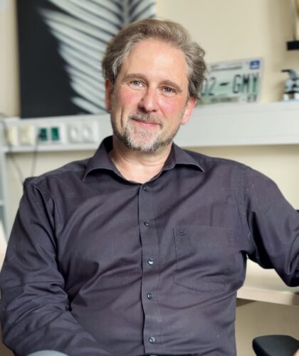 Prof. Harald Plank, ein Mann mittleren Alters mit hellem Haar und Bart, trägt ein dunkles Button-up-Hemd, sitzt in einem Büro. Er lächelt leicht und stützt einen Arm auf den Schreibtisch; hinter ihm sind Regale, ein Nummernschild und Kunstwerke zu sehen.