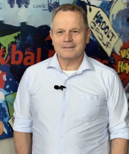 Prof. Harald Plank, im hellblauen Hemd, steht selbstbewusst vor einer bunten Graffiti-Wand mit Begriffen aus der Logistik und Lieferkette, ein Mikrofon am Hemd befestigt, in die Kamera.