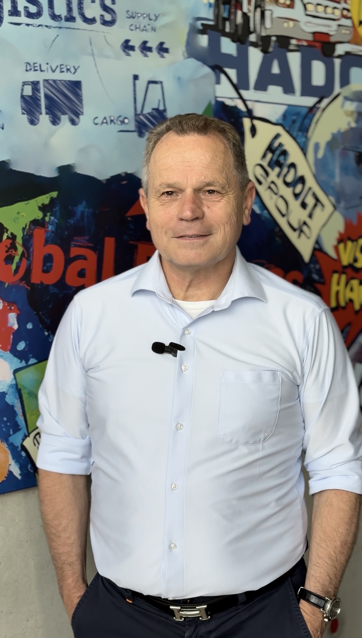 Prof. Harald Plank, im hellblauen Hemd, steht selbstbewusst vor einer bunten Graffiti-Wand mit Begriffen aus der Logistik und Lieferkette, ein Mikrofon am Hemd befestigt, in die Kamera.