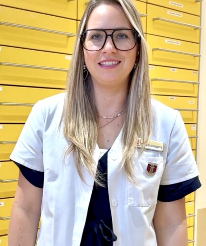 Eine Frau mit langen blonden Haaren und Brille lächelt, während sie einen weißen Laborkittel trägt. Sie steht vor einer Wand mit leuchtend gelben Schubladen.