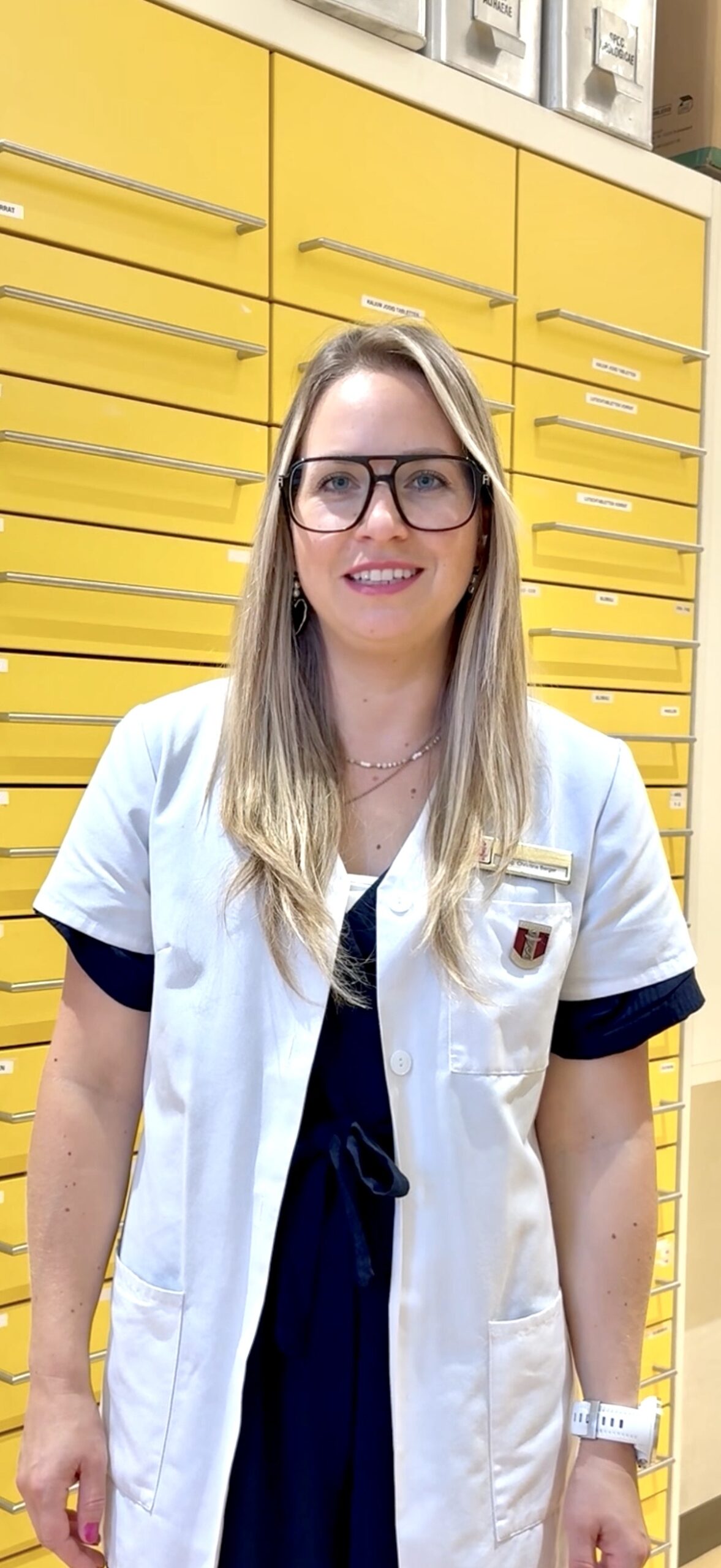 Eine Frau mit langen blonden Haaren und Brille lächelt, während sie einen weißen Laborkittel trägt. Sie steht vor einer Wand mit leuchtend gelben Schubladen.