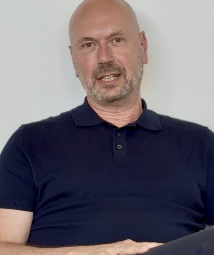 Ein Mann mittleren Alters mit Glatze und gestutztem Bart, der ein marineblaues Poloshirt trägt, sitzt vor einem schlichten hellen Hintergrund. Er blickt mit neutraler Miene in die Kamera und ist bereit für "3 Fragen an MCG-Vorstand Martin Ullrich".