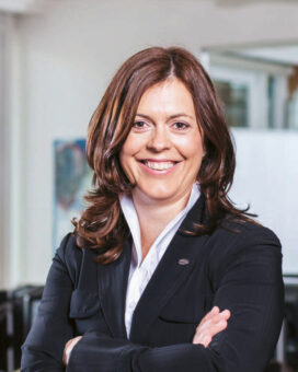 Eine Frau in einem schwarzen Anzug steht selbstbewusst mit verschränkten Armen in einem modernen, hellen Büro. Im Hintergrund arbeiten mehrere Personen an Schreibtischen, und um den Arbeitsplatz herum sind Grünpflanzen zu sehen.
