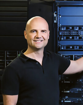 Ein lächelnder Mann in einem schwarzen Polohemd steht in einem Serverraum und stützt sich mit einer Hand auf ein hohes Serverregal, das mit Netzwerkgeräten und Servern gefüllt ist.
