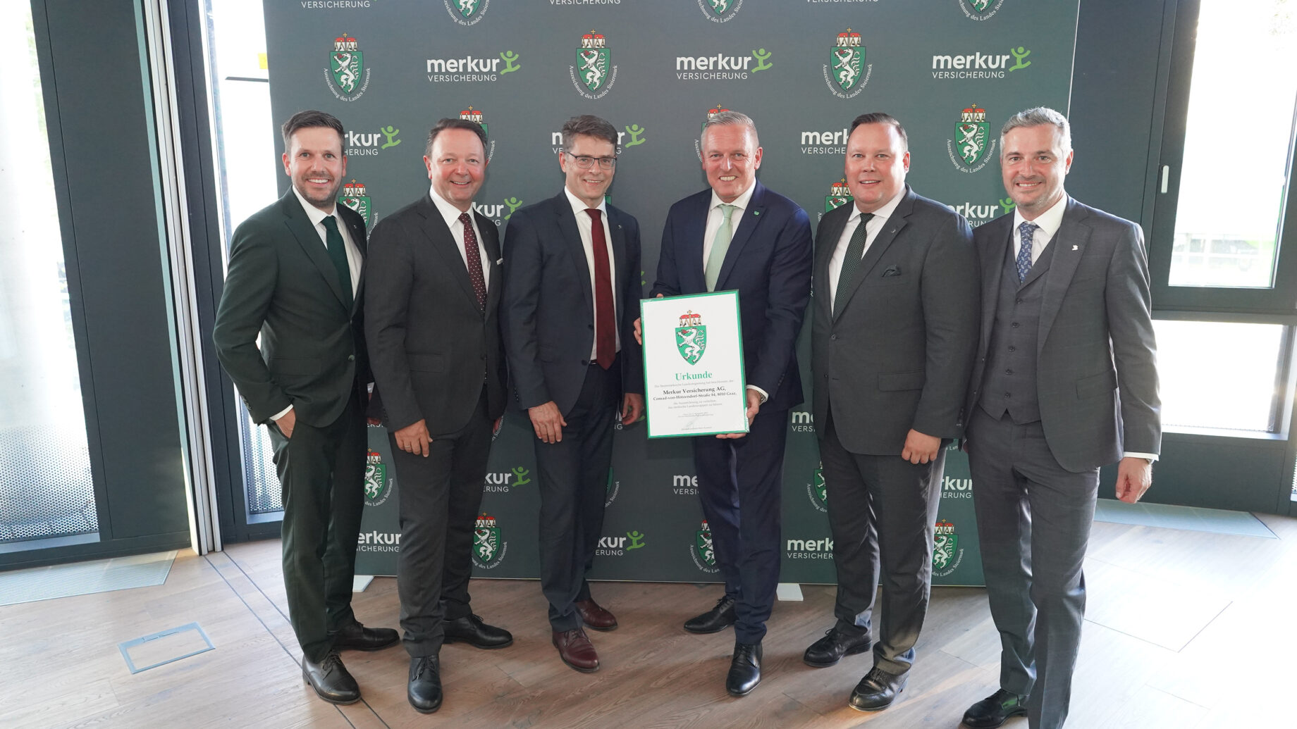 Sechs Männer in Anzügen stehen lächelnd vor einer Kulisse mit "merkur" und einem Wappenlogo bei der langen Tafel Graz. Der Mann in der Mitte hält eine gerahmte Urkunde in der Hand, während sie gemeinsam in einem Innenraum auf einem Holzboden posieren.