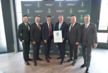 Sechs Männer in Anzügen stehen lächelnd vor einer Kulisse mit "merkur" und einem Wappenlogo bei der langen Tafel Graz. Der Mann in der Mitte hält eine gerahmte Urkunde in der Hand, während sie gemeinsam in einem Innenraum auf einem Holzboden posieren.