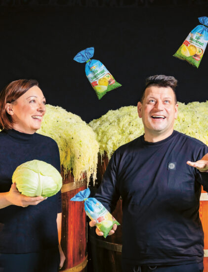 Eine Frau, die einen Kohl in der Hand hält, und ein Mann, der verpacktes Sauerkraut schwenkt, stehen lächelnd vor großen Holzfässern, die mit geschreddertem Kohl gefüllt sind. Drei Sauerkrautpakete schweben über ihnen.