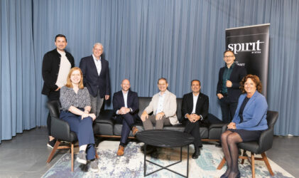 Acht Personen in Geschäftskleidung posieren für ein Gruppenfoto vor einem blauen Vorhang. Vier von ihnen sitzen auf einem Sofa und vier stehen hinter ihnen. Rechts ist ein schwarzes Schild mit der weißen Aufschrift "spirit" zu sehen.
