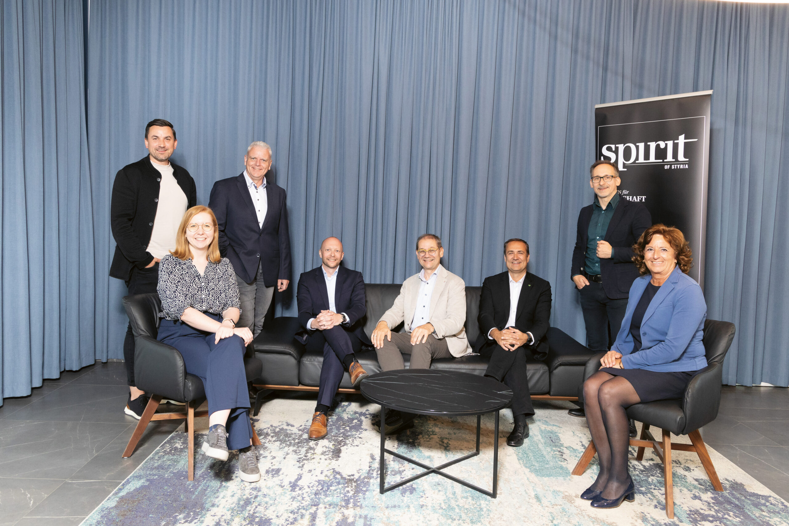 Acht Personen in Geschäftskleidung posieren für ein Gruppenfoto vor einem blauen Vorhang. Vier von ihnen sitzen auf einem Sofa und vier stehen hinter ihnen. Rechts ist ein schwarzes Schild mit der weißen Aufschrift "spirit" zu sehen.