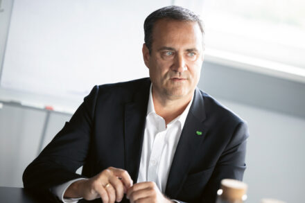 Ein Mann in einem dunklen Anzug und weißem Hemd sitzt an einem Tisch und schaut leicht nach links. Er scheint ein Gespräch zu führen und hat einen neutralen Gesichtsausdruck. Im Hintergrund sind ein Whiteboard und ein sonnendurchflutetes Fenster zu sehen.