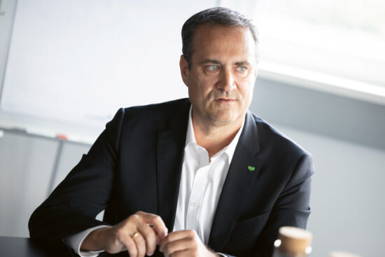 Ein Mann in einem dunklen Anzug und weißem Hemd sitzt an einem Tisch und schaut leicht nach links. Er scheint ein Gespräch zu führen und hat einen neutralen Gesichtsausdruck. Im Hintergrund sind ein Whiteboard und ein sonnendurchflutetes Fenster zu sehen.