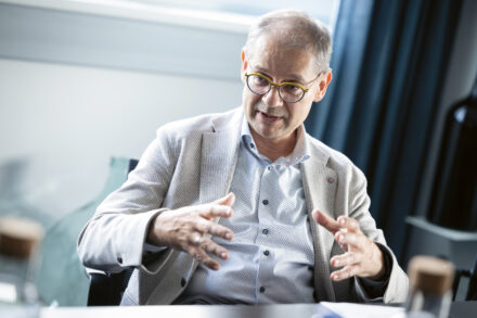 Ein Mann, der eine Brille und einen hellen Blazer trägt, gestikuliert mit den Händen, während er spricht. Er sitzt in einem Innenraum an einem Tisch mit verschwommenen Flaschen im Vordergrund und einem Fenster im Hintergrund.