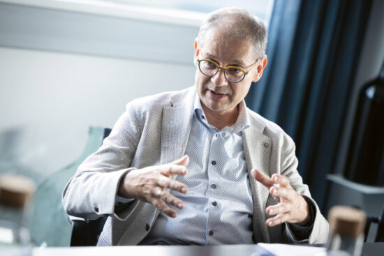 Ein Mann, der eine Brille und einen hellen Blazer trägt, gestikuliert mit den Händen, während er spricht. Er sitzt in einem Innenraum an einem Tisch mit verschwommenen Flaschen im Vordergrund und einem Fenster im Hintergrund.