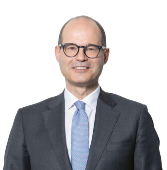 Ein Mann in einem dunklen Anzug, mit hellblauer Krawatte und Brille steht lächelnd vor einem schlichten weißen Hintergrund und hat die Hände vor sich verschränkt.