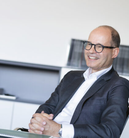 Ein Mann im Anzug und mit Brille sitzt an einem Schreibtisch und lächelt mit gefalteten Händen. Im Hintergrund sind Regale und Akten zu sehen, was auf eine Büroumgebung hindeutet.