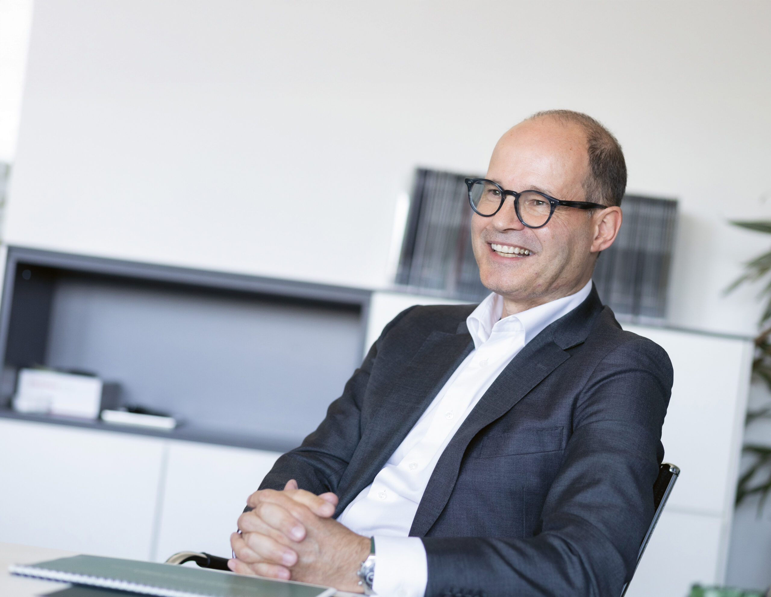 Ein Mann im Anzug und mit Brille sitzt an einem Schreibtisch und lächelt mit gefalteten Händen. Im Hintergrund sind Regale und Akten zu sehen, was auf eine Büroumgebung hindeutet.
