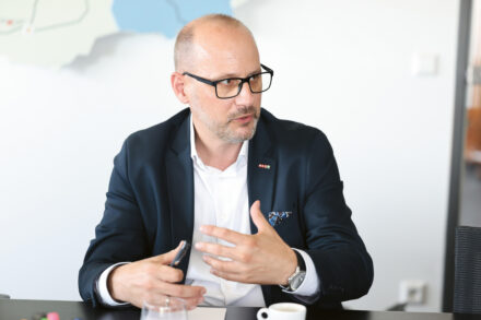 Ein Mann mit Brille und Bart, bekleidet mit einem marineblauen Blazer und einem weißen Hemd, sitzt an einem Tisch und spricht und gestikuliert mit den Händen. Im Hintergrund sind eine Kaffeetasse, Papiere und eine Landkarte an der Wand zu sehen.