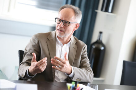 Ein Mann mit Brille und beigem Blazer gestikuliert mit den Händen, während er an einem Tisch mit Büromaterial in einem hellen, modernen Raum spricht.