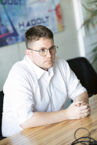 Ein Mann mit kurzen Haaren und Brille, der ein weißes Hemd trägt, sitzt an einem Holztisch, hat die Hände gefaltet und schaut aufmerksam. Im Hintergrund ist ein buntes, unscharfes Plakat zu sehen.