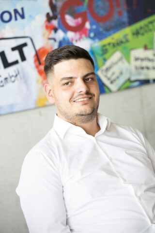 Ein Mann mit kurzen dunklen Haaren und gestutztem Bart, der ein weißes Button-up-Hemd trägt, lächelt, während er vor einer bunten Wand im Graffiti-Stil sitzt.