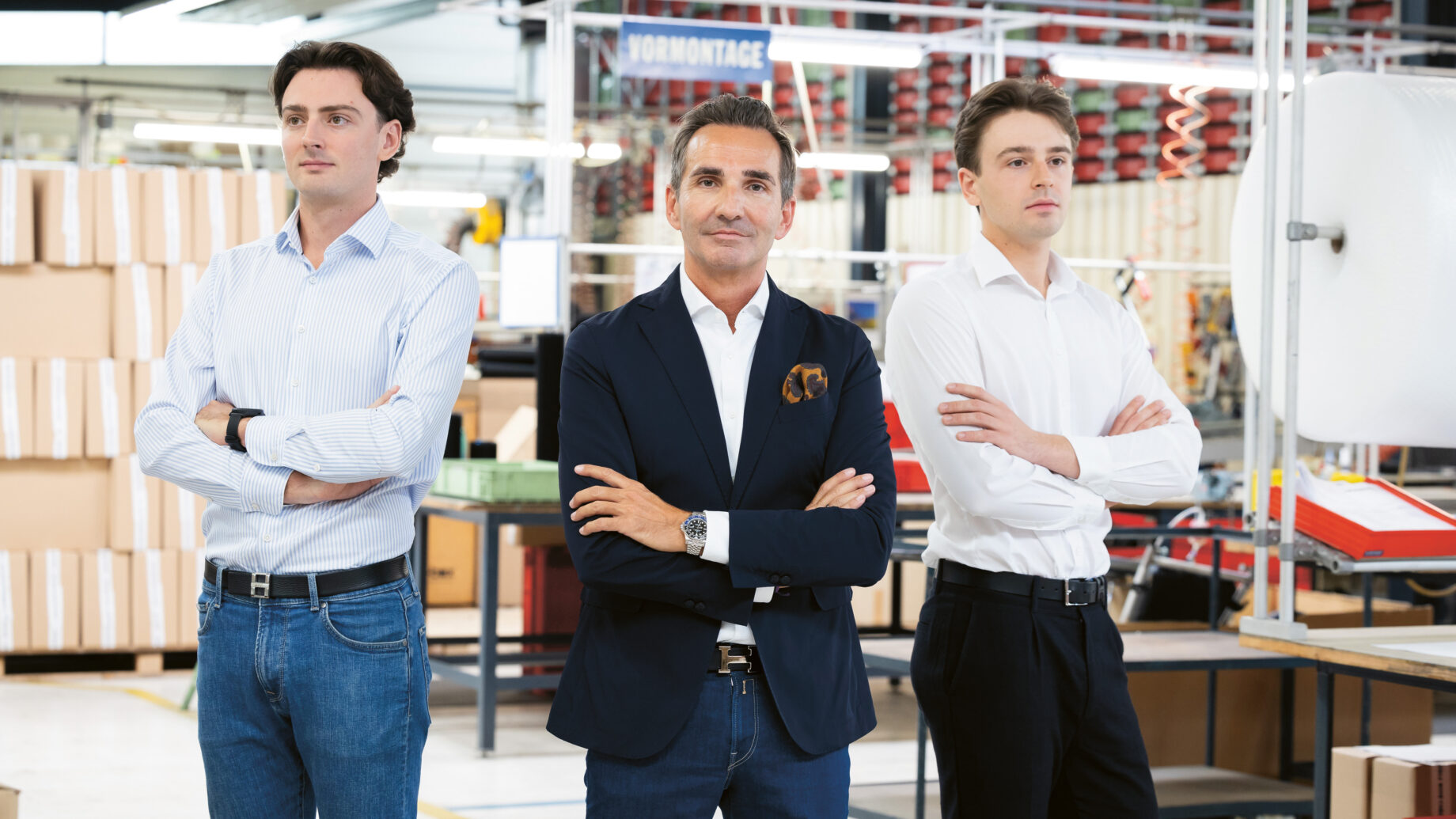 Drei Männer in Geschäftskleidung stehen mit verschränkten Armen in einer modernen, hell erleuchteten Fabrik oder Lagerhalle, umgeben von Regalen, Kisten und Industrieanlagen.