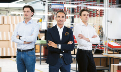 Drei Männer in Geschäftskleidung stehen mit verschränkten Armen in einer modernen, hell erleuchteten Fabrik oder Lagerhalle, umgeben von Regalen, Kisten und Industrieanlagen.