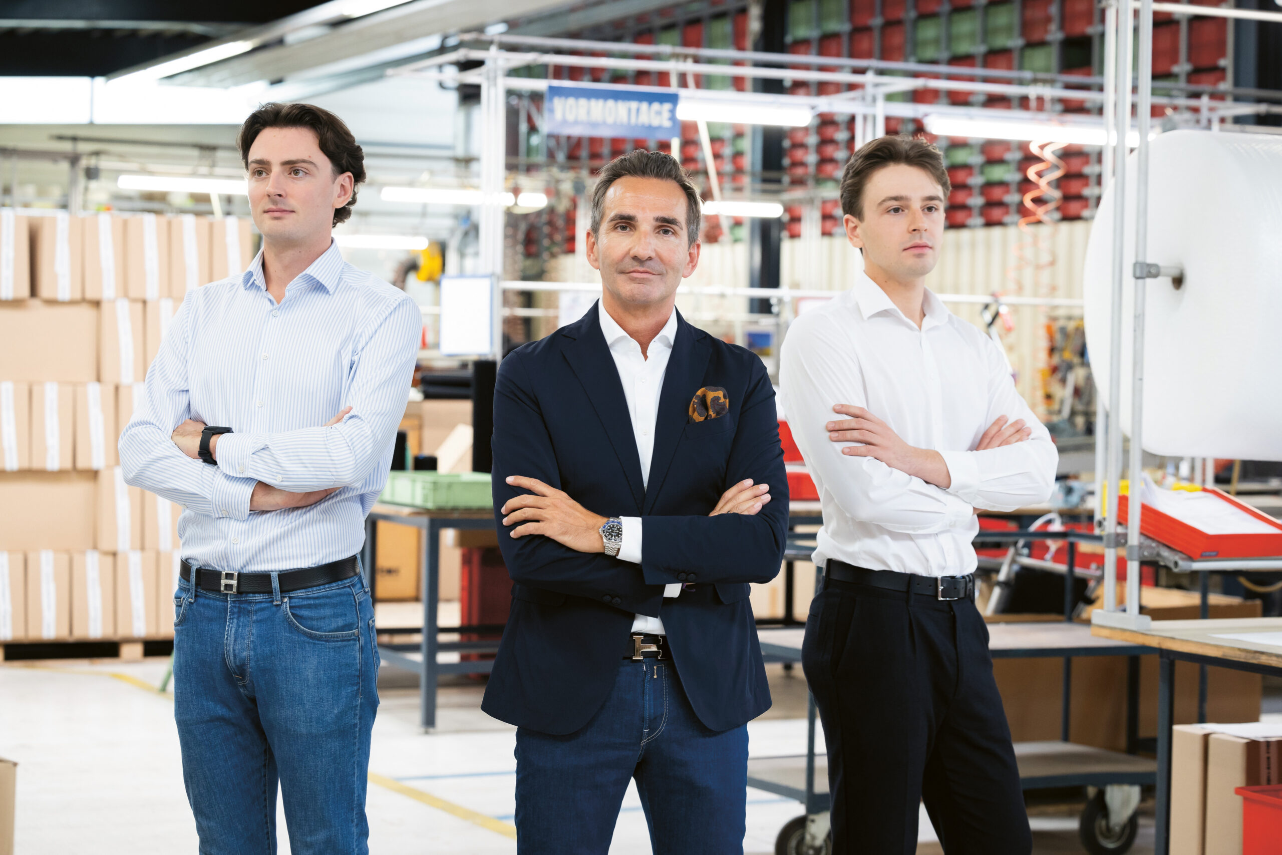 Drei Männer in Geschäftskleidung stehen mit verschränkten Armen in einer modernen, hell erleuchteten Fabrik oder Lagerhalle, umgeben von Regalen, Kisten und Industrieanlagen.