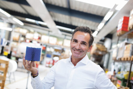 Ein lächelnder Mann in einem weißen Hemd hält eine blaue, stachelige Bürste in einem hell erleuchteten Lagerhaus oder einer Werkstatt hoch, wobei im Hintergrund Regale und Kisten zu sehen sind.