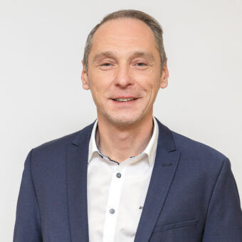 Ein Mann in einem marineblauen Blazer und einem weißen Hemd lächelt leicht, während er vor einem schlichten weißen Hintergrund steht.