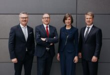 Vier Geschäftsleute, drei Männer und eine Frau, stehen in formeller Kleidung nebeneinander vor einer modernen grauen Wand und lächeln in die Kamera - bereit für die lange Tafel Graz.