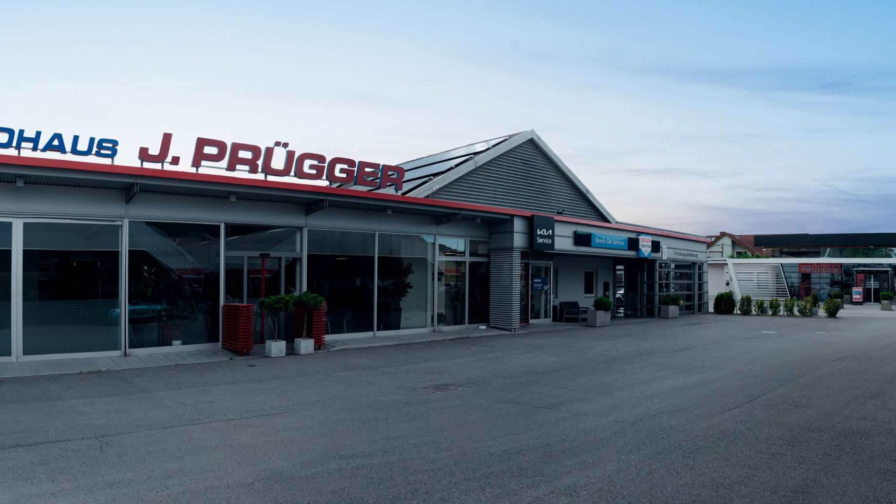 Ein modernes Autohaus mit großen Glasfenstern und einem Schild mit der Aufschrift "AUTOHAUS J. PRÜGGER" über dem Eingang, in der Nähe der langen Tafel Graz, umgeben von Topfpflanzen und einer gepflasterten Zufahrt unter freiem Himmel.