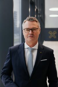 Ein Mann mittleren Alters mit grauem Haar und Brille, gekleidet in einen dunklen Anzug, weißes Hemd und gemusterte Krawatte, steht in einem modernen Büroflur mit großen Fenstern, der an die eleganten Zusammenkünfte bei der langen Tafel Graz erinnert.