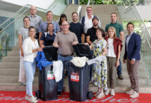 Eine Gruppe von 14 lächelnden Erwachsenen posiert drinnen vor einer Treppe und steht um zwei schwarze Mülltonnen mit Stoffen, die auf eine Investition in ihrem Regionalmarkt Steiermark hinweisen und ein Engagement für Gemeinschaft und Nachhaltigkeit zeigen.