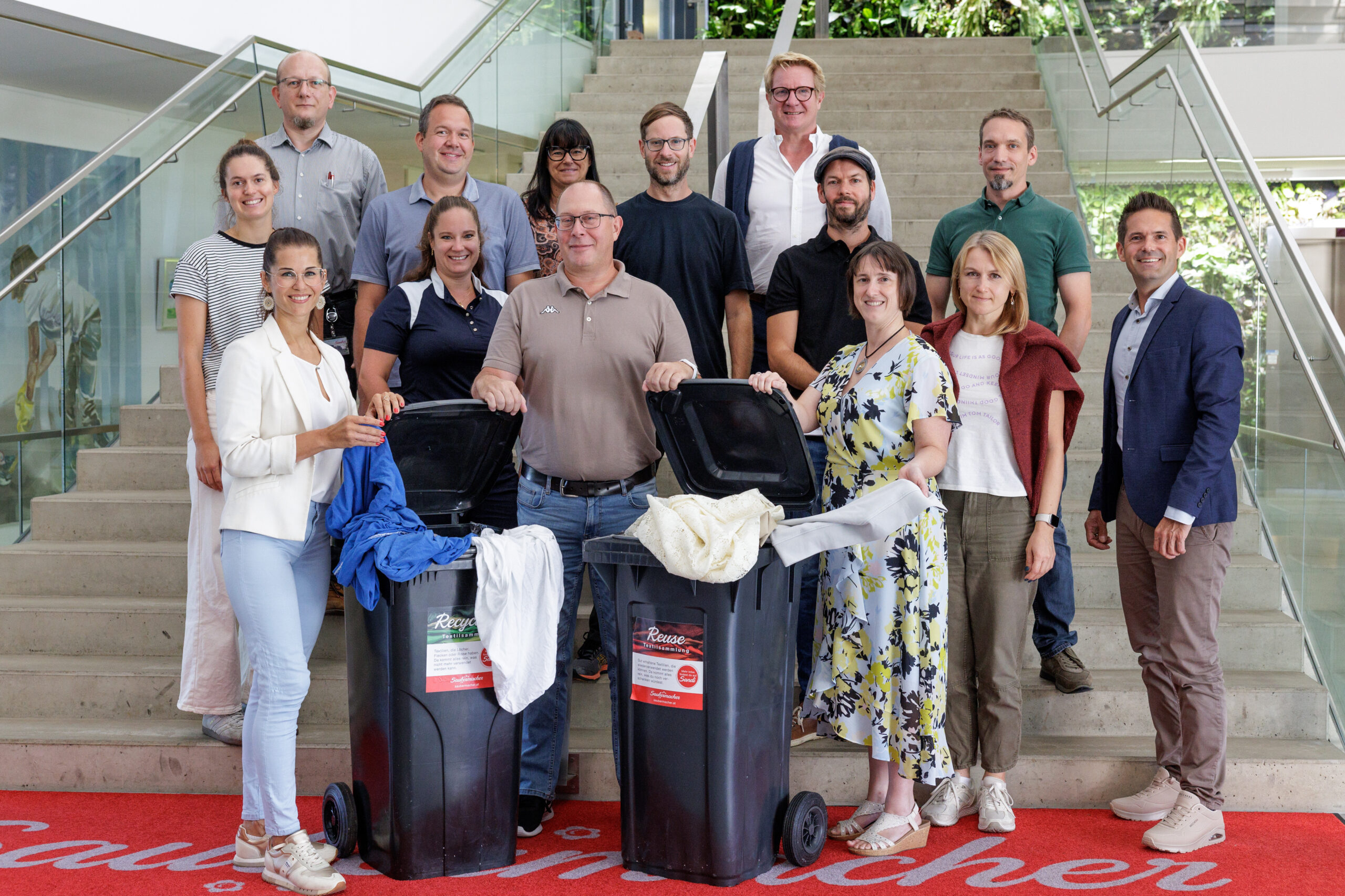 Eine Gruppe von 14 lächelnden Erwachsenen posiert drinnen vor einer Treppe und steht um zwei schwarze Mülltonnen mit Stoffen, die auf eine Investition in ihrem Regionalmarkt Steiermark hinweisen und ein Engagement für Gemeinschaft und Nachhaltigkeit zeigen.