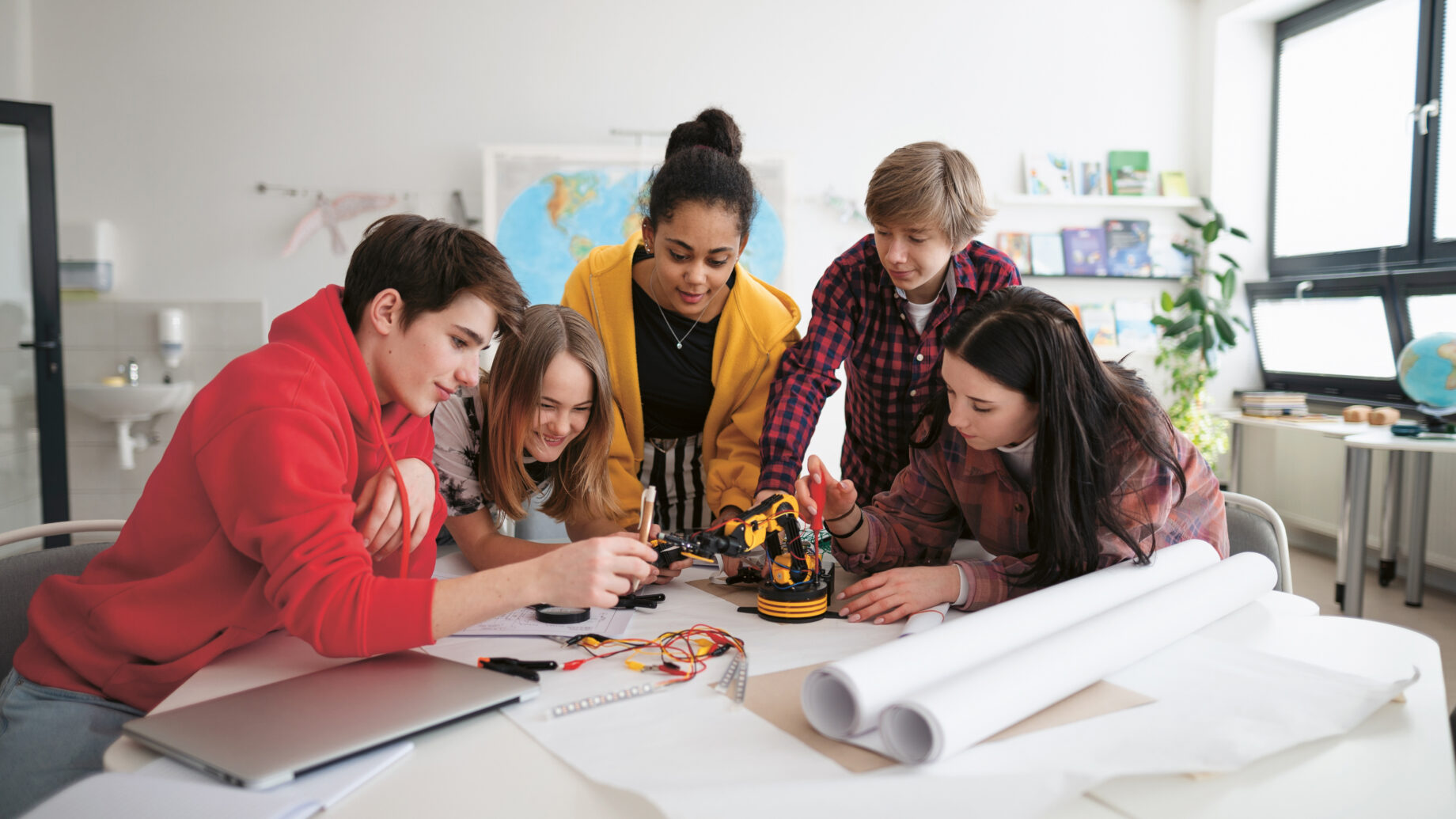 Fünf Jugendliche versammeln sich um einen Tisch und arbeiten gemeinsam an einem kleinen Projekt mit einem Roboterarm. Sie lächeln und arbeiten engagiert, mit Plänen, Kabeln und einem Laptop auf dem Tisch in einem hellen Klassenzimmer.