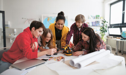 Fünf Jugendliche versammeln sich um einen Tisch und arbeiten gemeinsam an einem kleinen Projekt mit einem Roboterarm. Sie lächeln und arbeiten engagiert, mit Plänen, Kabeln und einem Laptop auf dem Tisch in einem hellen Klassenzimmer.