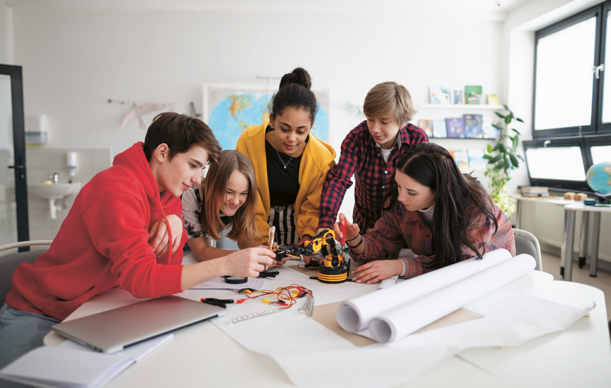 Fünf Jugendliche versammeln sich um einen Tisch und arbeiten gemeinsam an einem kleinen Projekt mit einem Roboterarm. Sie lächeln und arbeiten engagiert, mit Plänen, Kabeln und einem Laptop auf dem Tisch in einem hellen Klassenzimmer.