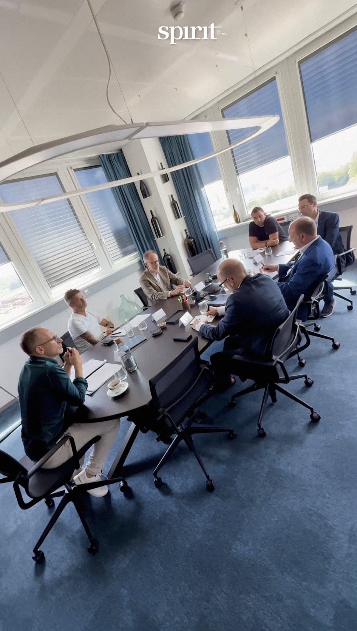 Sechs Personen sitzen um einen großen Konferenztisch in einem modernen Büro und diskutieren. Laptops, Papiere und Getränke stehen auf dem Tisch, während natürliches Licht hereinströmt. "Apothekerin des Jahres: Alexandra Fuchsbichler gratuliert" ist am oberen Rand zu lesen.
