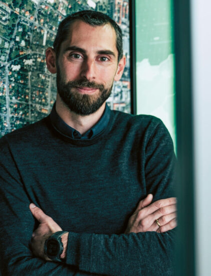 Ein Mann mit kurzen Haaren und Bart, der einen dunklen Pullover und eine Smartwatch trägt, steht mit verschränkten Armen vor einem großen Karten- oder Luftbild-Wanddisplay.