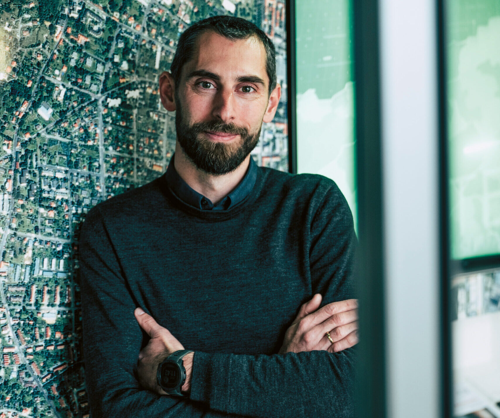 Ein Mann mit kurzen Haaren und Bart, der einen dunklen Pullover und eine Smartwatch trägt, steht mit verschränkten Armen vor einem großen Karten- oder Luftbild-Wanddisplay.