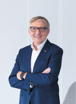 Ein lächelnder Mann in einem marineblauen Anzug und mit Brille steht mit verschränkten Armen vor einer modernen Wand mit dem verpixelten Graustufenporträt eines anderen Mannes.