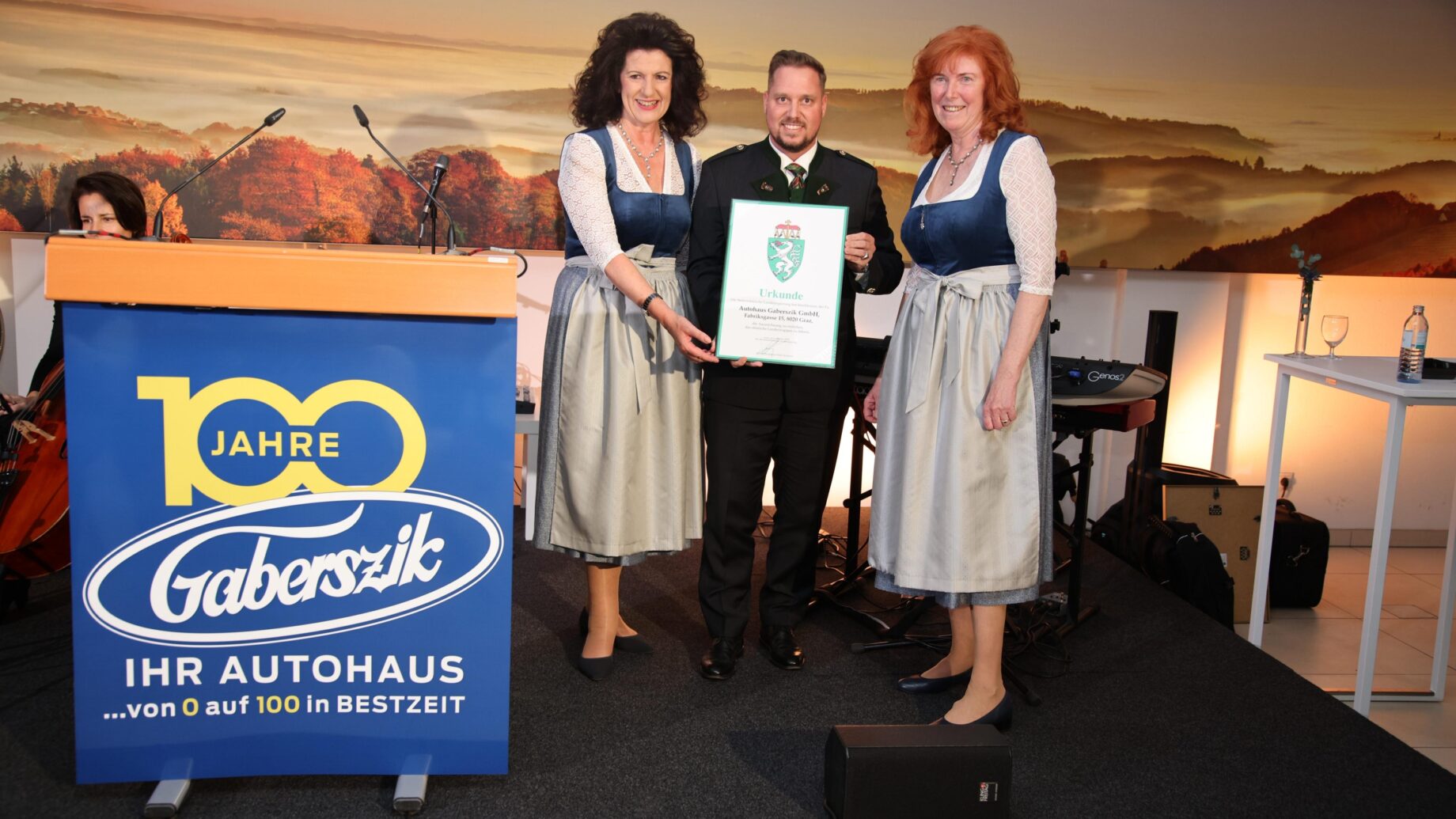 Drei Personen in bayerischer Tracht stehen auf einer Bühne bei den Holz steirischen Tischlertagen; der Mann in der Mitte hält eine Urkunde. Ein Podium mit der Aufschrift "100 Jahre Gaberszik" und ein landschaftlicher Hintergrund sind zu sehen.